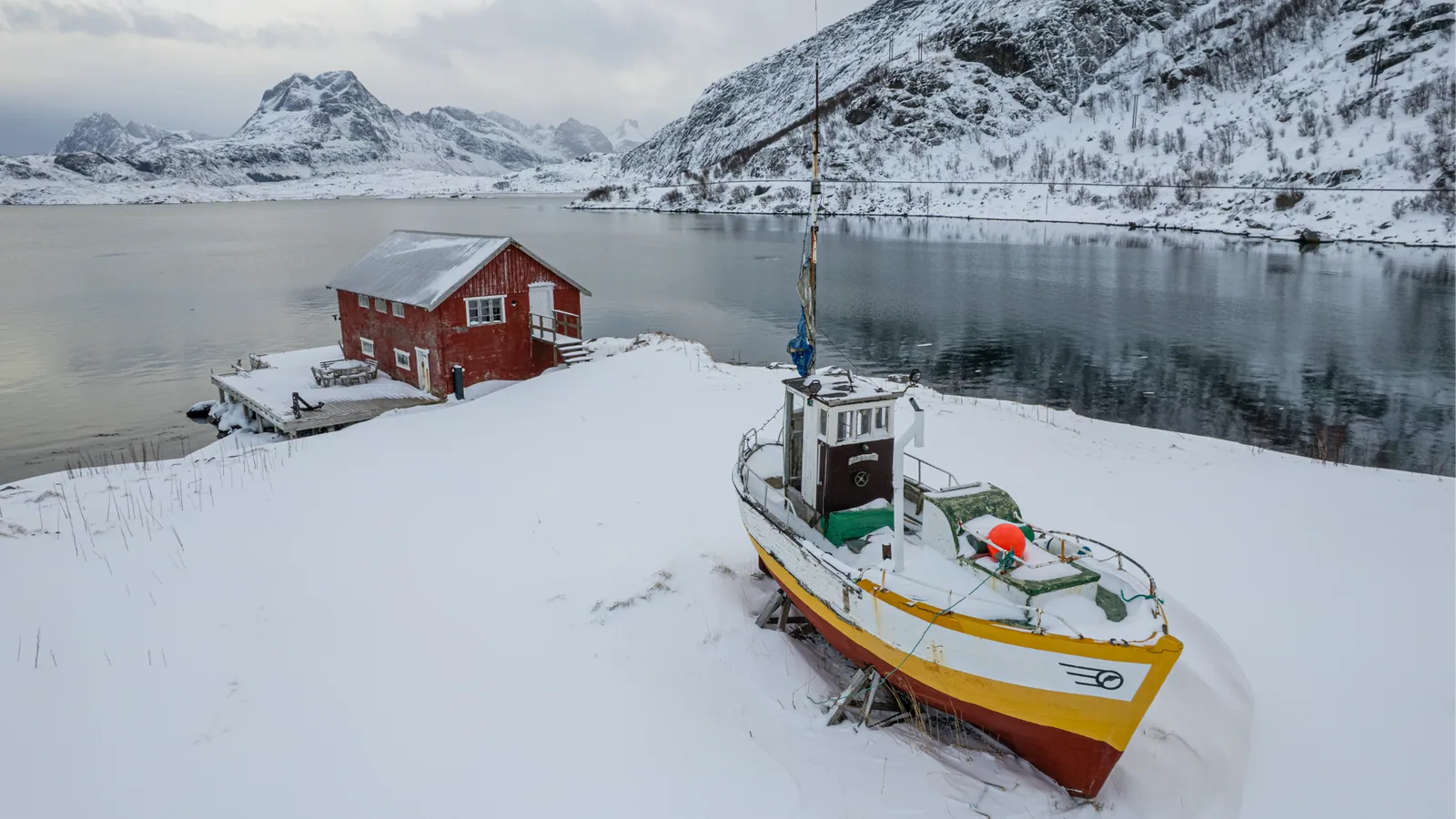 A red cabin and a fishing boat beside a snowy fjord in the Lofoten Islands
