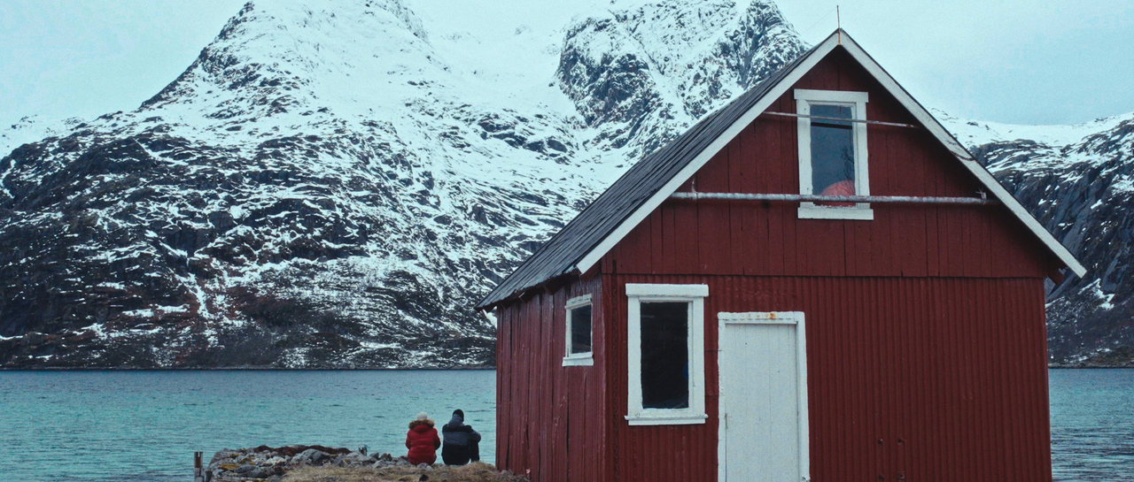Two people sit beside a red cabin beneath snow-covered mountains